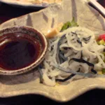 Un petit bol de ponzu sombre posé à côté de fines lanières de peau de fugu et d'oignon nouveau, dans un izakaya de Tokyo