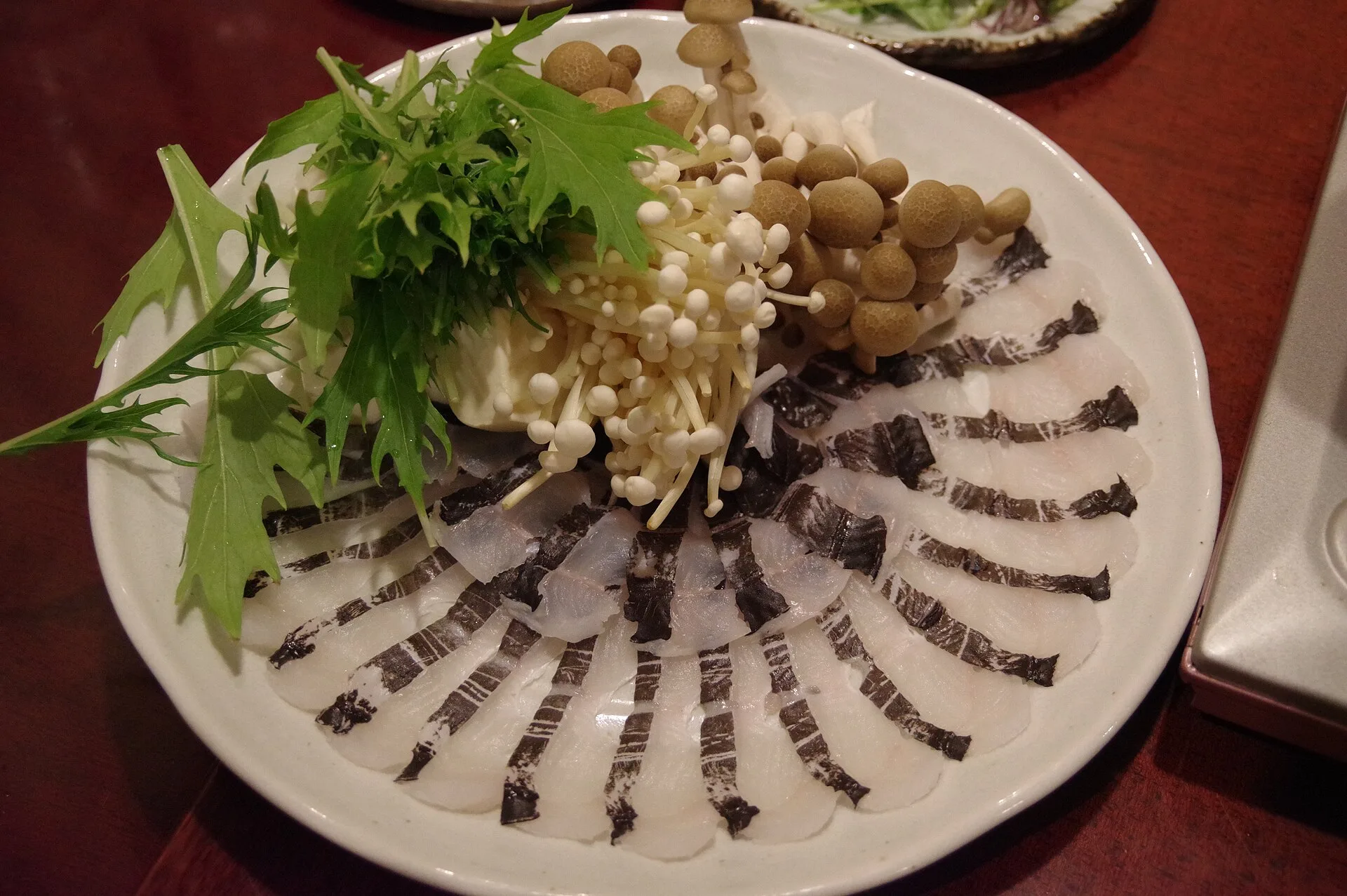 Une assiette de fines tranches de congre crues disposées en éventail, avec des champignons enoki, des shimeji et des feuilles de mizuna
