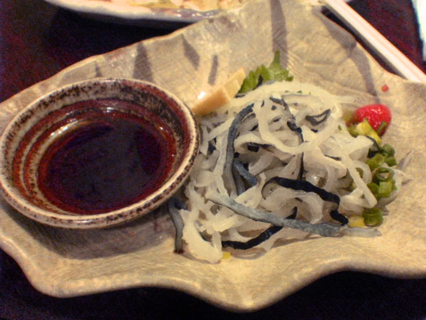 Un petit bol de ponzu sombre posé à côté de fines lanières de peau de fugu et d'oignon nouveau, dans un izakaya de Tokyo