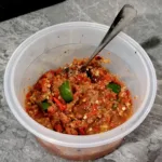 Un pot de sambal belacan maison, une pâte rouge grossièrement pilée avec des quartiers d'agrume vert, une cuillère plantée dedans