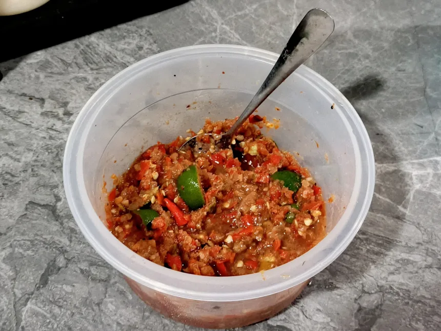 Un pot de sambal belacan maison, une pâte rouge grossièrement pilée avec des quartiers d'agrume vert, une cuillère plantée dedans