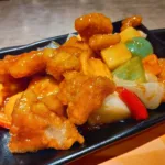 Des morceaux de porc frits nappés d'une sauce orangée brillante, avec des dés d'ananas, de poivron et d'oignon, sur une assiette noire