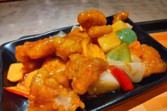 Des morceaux de porc frits nappés d'une sauce orangée brillante, avec des dés d'ananas, de poivron et d'oignon, sur une assiette noire