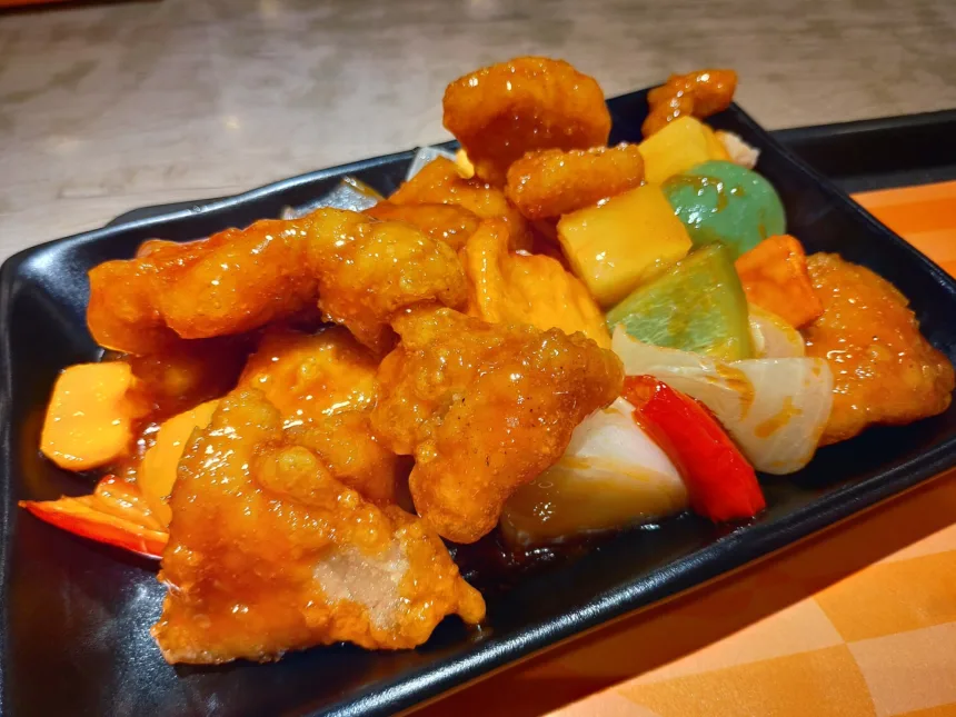 Des morceaux de porc frits nappés d'une sauce orangée brillante, avec des dés d'ananas, de poivron et d'oignon, sur une assiette noire