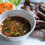 Un petit bol de sauce brun rougeâtre parsemée de flocons de piment, de grains de riz grillé et d'herbes hachées
