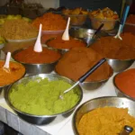 Un étal de marché couvert de bassines en inox remplies de pâtes de curry rouges, orange, brunes et vertes, chacune avec sa cuillère