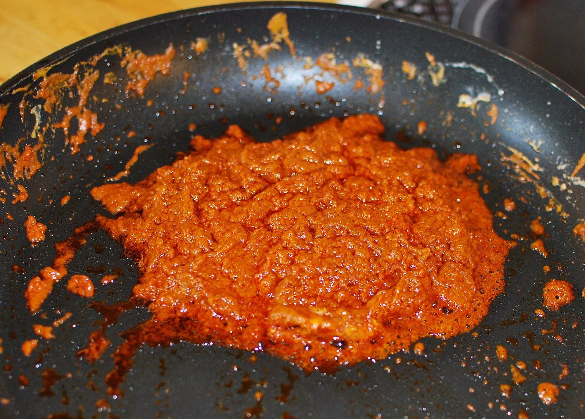 Une pâte de curry rouge orangé en train de frire dans une poêle noire, entourée de gouttes d'huile