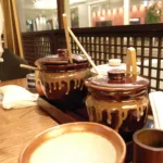 Deux pots de sauce en grès brun, louche en bois plantée dans le couvercle, posés sur la table d'un restaurant de Shinjuku, avec un petit mortier strié au premier plan