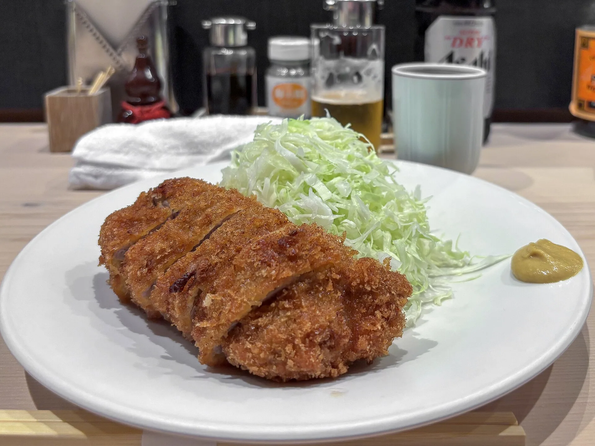 Sauce tonkatsu : la recette japonaise et ses équivalents 8 Un tonkatsu doré coupé en tranches sur une assiette blanche, avec un dôme de chou cru émincé et une noisette de moutarde jaune, devant un comptoir garni de bouteilles