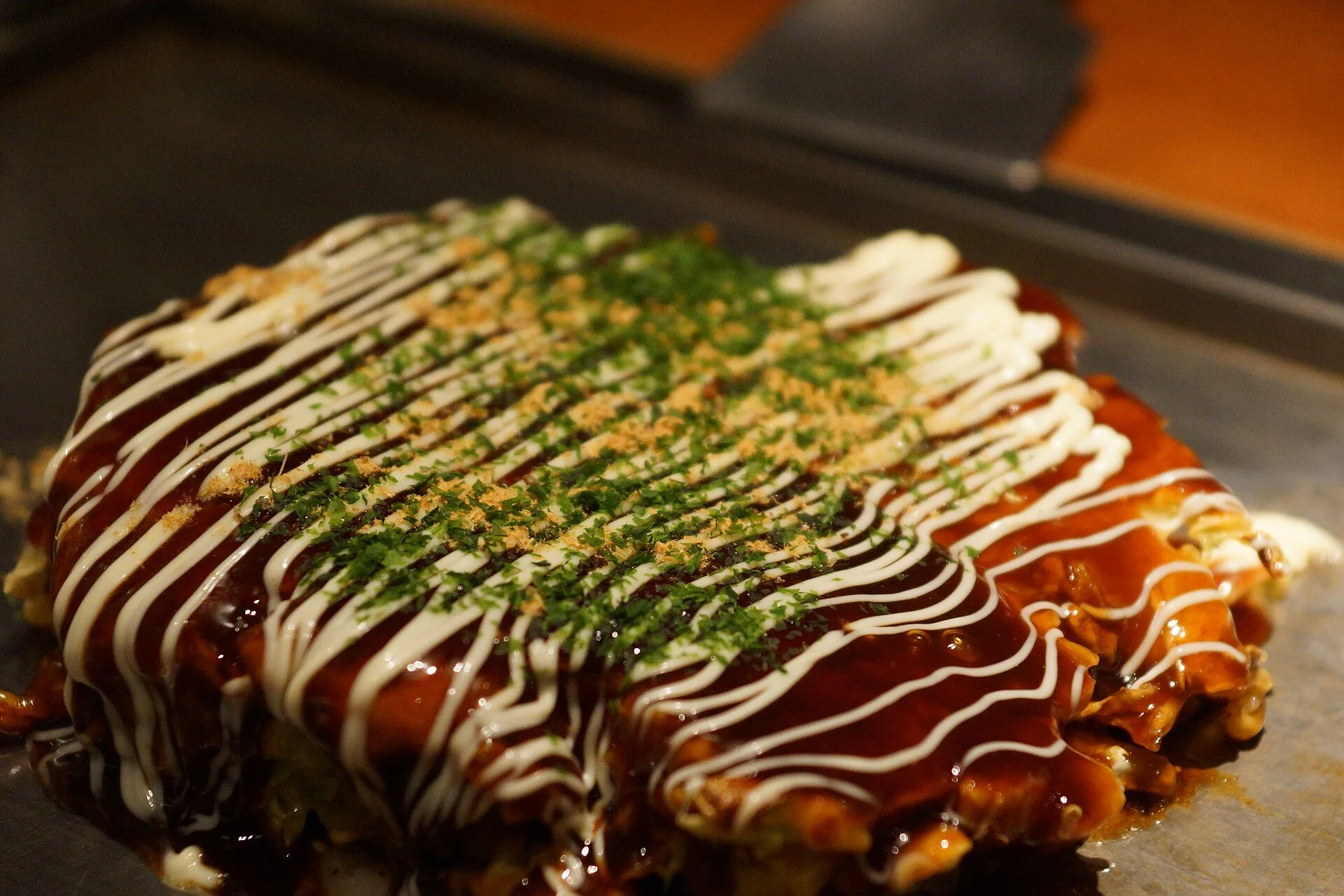 Sauce tonkatsu : la recette japonaise et ses équivalents 9 Une galette d'okonomiyaki nappée de sauce brune brillante, striée de mayonnaise et saupoudrée d'algue verte et de bonite, sur une plaque chauffante