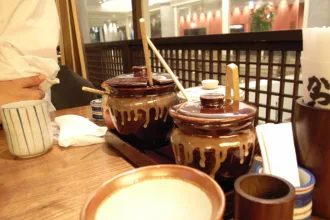 Deux pots de sauce en grès brun, louche en bois plantée dans le couvercle, posés sur la table d'un restaurant de Shinjuku, avec un petit mortier strié au premier plan
