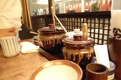 Deux pots de sauce en grès brun, louche en bois plantée dans le couvercle, posés sur la table d'un restaurant de Shinjuku, avec un petit mortier strié au premier plan
