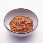 Un bol de céramique blanche cerclé de brun, rempli d'une pâte épaisse brun orangé parsemée de graines de sésame et de petits morceaux de piment vert