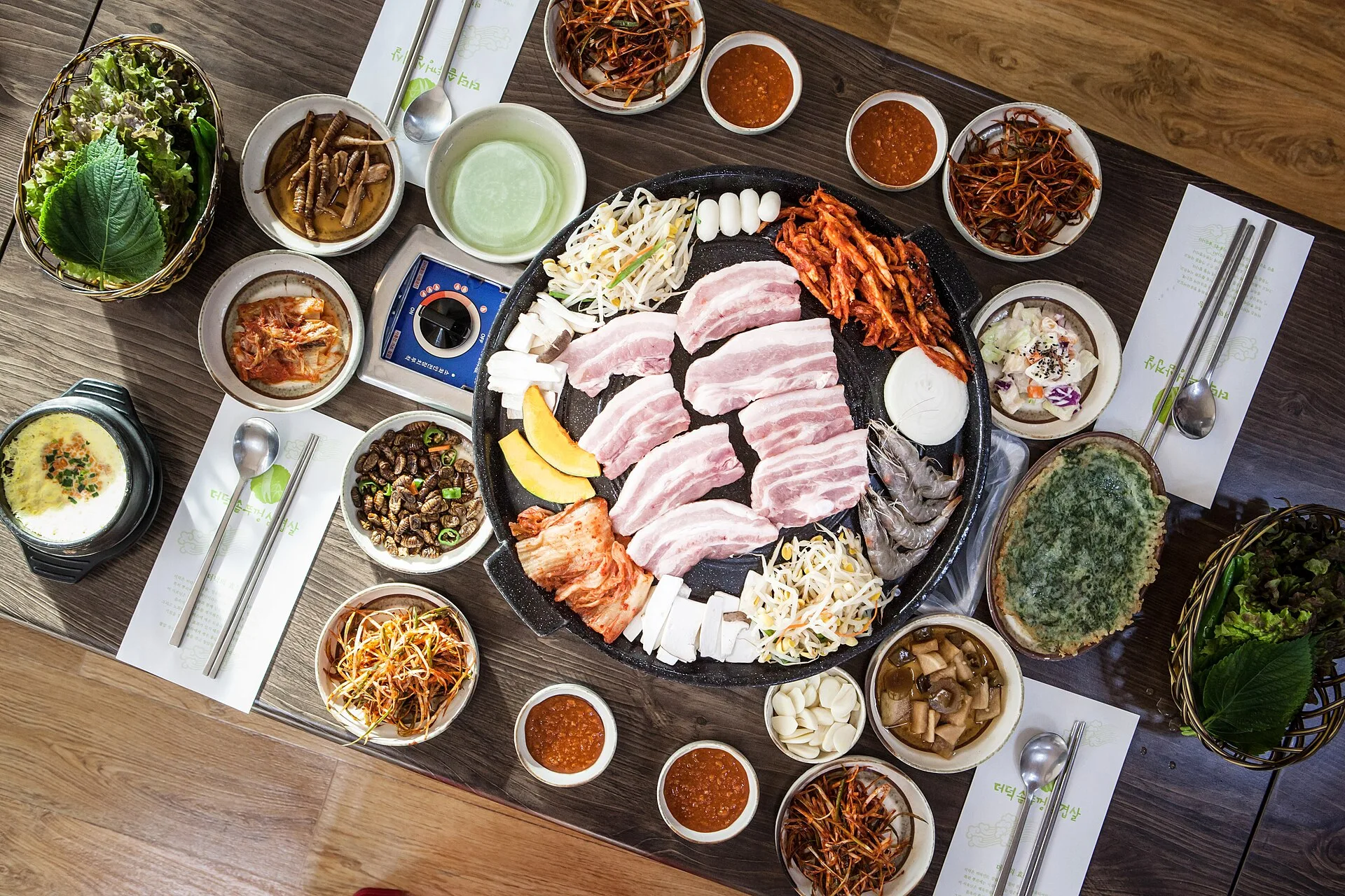 Ssamjang : la sauce du barbecue coréen, recette et ratio 8 Vue du dessus d'une table de restaurant coréen : tranches de poitrine de porc crue sur une plaque ronde, entourées de petits plats d'accompagnement, de ramequins de sauce orangée et de deux paniers de salade et de feuilles de pérille
