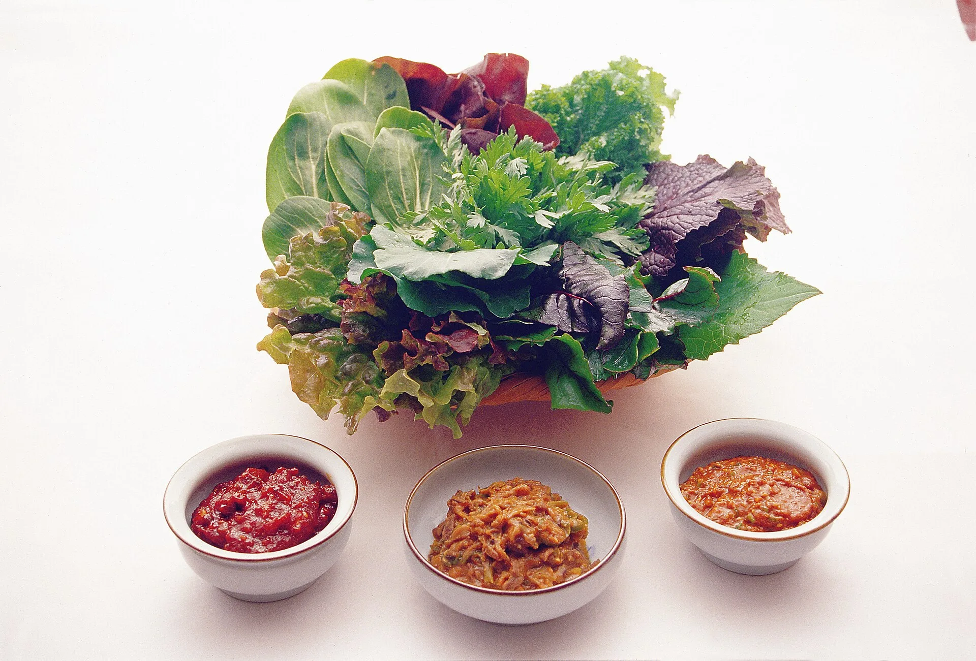 Ssamjang : la sauce du barbecue coréen, recette et ratio 9 Un panier débordant de feuilles de salade verte et rouge, de feuilles de pérille et de chrysanthème, devant trois petits bols de pâtes de couleurs différentes, du rouge vif au brun